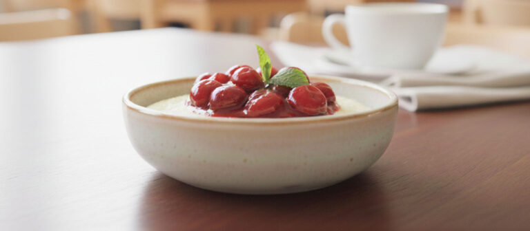 Eine helle, strukturierte Keramikschale, gefüllt mit cremigem Grießbrei, der mit glänzenden, roten Kirschen und einem frischen Minzblatt garniert ist.