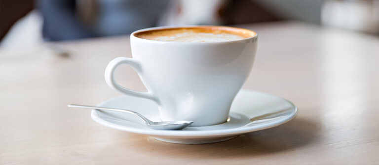 Eine weiße Tasse mit einem Kafee mit Milchschaum. Die Tasse steht auf einer passenden weißen Untertasse, daneben liegt ein kleiner silberner Löffel. Der Hintergrund ist unscharf.