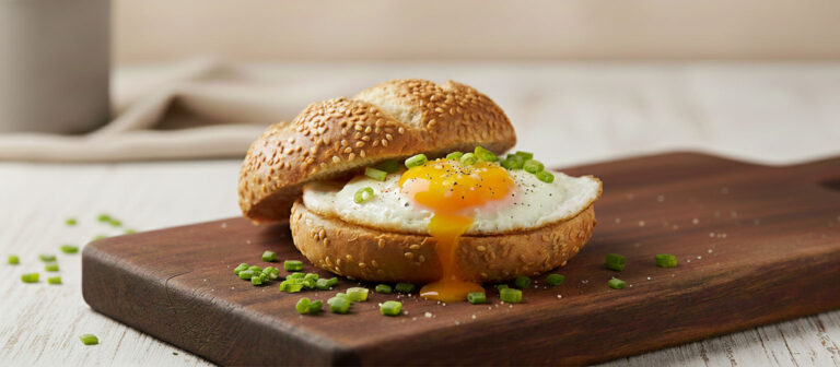Ein geöffnetes Sesambrötchen mit einem Spiegelei in der Mitte, dessen flüssiges Eigelb sanft über das Brötchen läuft. Das Ei ist mit Pfeffer bestreut und das Gericht ist mit frischem Schnittlauch garniert. Es liegt auf einem dunklen Holzschneidebrett, auf dem ebenfalls Schnittlauch verstreut ist.